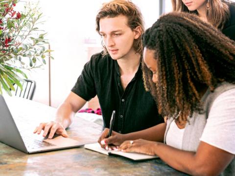 A man and two women reviewing a laptop screen and writing notes