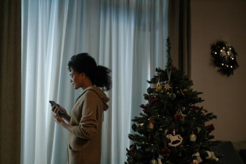 a woman stands by a curtain and a Christmas tree and looks sadly at her phone 