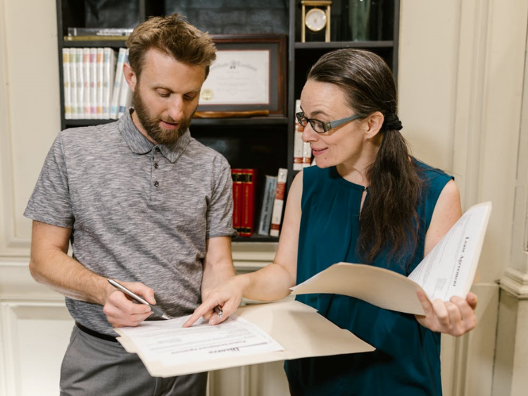 Woman pointing to a legal document, indicating where to sign to a man, who holds the document open in his hands.