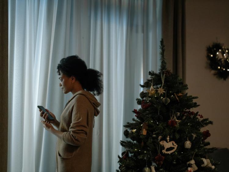 a woman stands by a curtain and a Christmas tree and looks sadly at her phone 